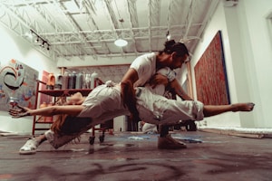 Two dancers are performing an intricate lift in an art studio with a high ceiling and various paintings and art supplies around. The woman is arched backward, balancing on the man's knee, while he supports her with his arms. The environment suggests a creative and dynamic energy.