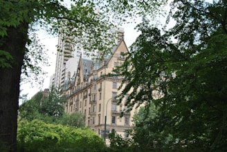 A historic building with a beige facade sits nestled among lush green trees, with the tops of modern high-rise buildings visible in the background. The architecture features pointed roofs and ornate detailing, giving it a classic, elegant appearance.
