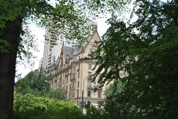 A historic building with a beige facade sits nestled among lush green trees, with the tops of modern high-rise buildings visible in the background. The architecture features pointed roofs and ornate detailing, giving it a classic, elegant appearance.