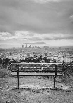 grayscale photo of bench near city buildings