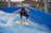 A person surfing on an artificial wave, performing a jump with a surfboard. The surfboard is labeled with 'HYPE' and other logos. The setting includes a blue wave pool with splashing water, surrounded by metal railings. In the background are people observing the activity, along with some floral plants.