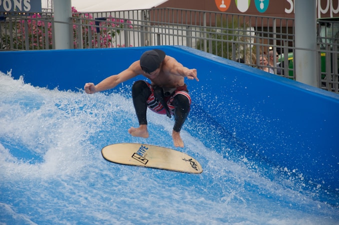 A person surfing on an artificial wave, performing a jump with a surfboard. The surfboard is labeled with 'HYPE' and other logos. The setting includes a blue wave pool with splashing water, surrounded by metal railings. In the background are people observing the activity, along with some floral plants.