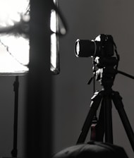 A professional camera is mounted on a tripod, positioned next to a large, soft light diffuser. The scene presents a typical photography studio setup, with focus on the camera equipment and lighting.