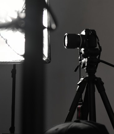 A professional camera is mounted on a tripod, positioned next to a large, soft light diffuser. The scene presents a typical photography studio setup, with focus on the camera equipment and lighting.