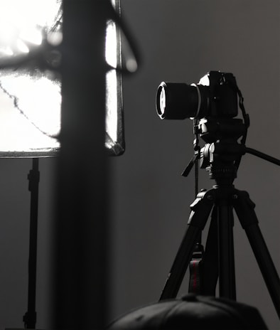 A professional camera is mounted on a tripod, positioned next to a large, soft light diffuser. The scene presents a typical photography studio setup, with focus on the camera equipment and lighting.