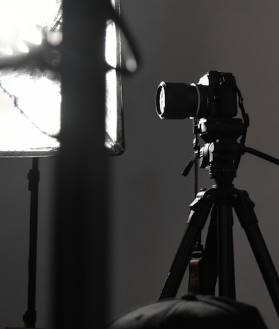 A professional camera is mounted on a tripod, positioned next to a large, soft light diffuser. The scene presents a typical photography studio setup, with focus on the camera equipment and lighting.