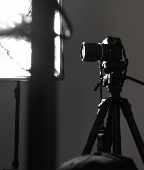 A professional camera is mounted on a tripod, positioned next to a large, soft light diffuser. The scene presents a typical photography studio setup, with focus on the camera equipment and lighting.