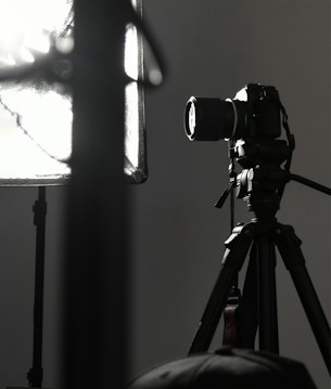 A professional camera is mounted on a tripod, positioned next to a large, soft light diffuser. The scene presents a typical photography studio setup, with focus on the camera equipment and lighting.