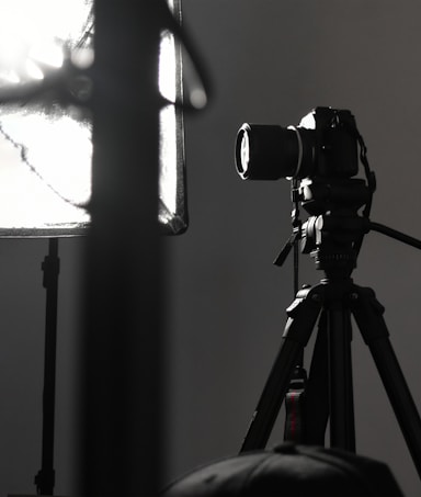A professional camera is mounted on a tripod, positioned next to a large, soft light diffuser. The scene presents a typical photography studio setup, with focus on the camera equipment and lighting.