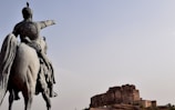 A large statue of a mounted, armored figure on a horse points towards an ancient fort in the distance under a clear sky.