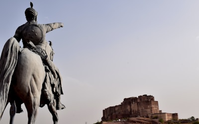 A large statue of a mounted, armored figure on a horse points towards an ancient fort in the distance under a clear sky.