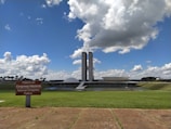 The photograph depicts a modern architectural building with two tall towers and a distinctive dome and bowl shape in the foreground. A lush green lawn stretches out in front of the structure under a bright blue sky filled with large, fluffy clouds. A sign in Portuguese and English reads 'Palácio do Congresso Nacional' and 'National Congress Palace'.