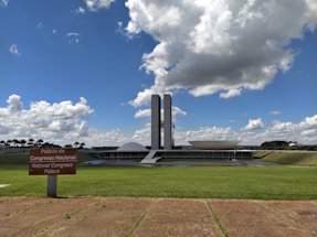 The photograph depicts a modern architectural building with two tall towers and a distinctive dome and bowl shape in the foreground. A lush green lawn stretches out in front of the structure under a bright blue sky filled with large, fluffy clouds. A sign in Portuguese and English reads 'Palácio do Congresso Nacional' and 'National Congress Palace'.