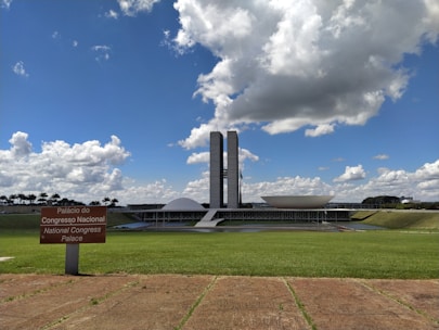 The photograph depicts a modern architectural building with two tall towers and a distinctive dome and bowl shape in the foreground. A lush green lawn stretches out in front of the structure under a bright blue sky filled with large, fluffy clouds. A sign in Portuguese and English reads 'Palácio do Congresso Nacional' and 'National Congress Palace'.