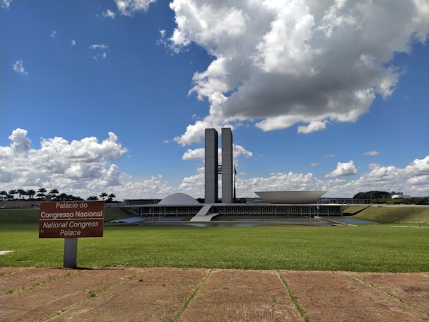 The photograph depicts a modern architectural building with two tall towers and a distinctive dome and bowl shape in the foreground. A lush green lawn stretches out in front of the structure under a bright blue sky filled with large, fluffy clouds. A sign in Portuguese and English reads 'Pal&aacute;cio do Congresso Nacional' and 'National Congress Palace'.