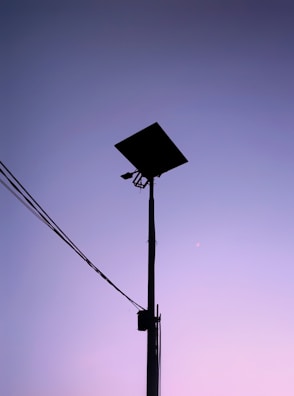 A silhouette of a tall street light with solar panels stands against a gradient sky transitioning from purple to pink. Electrical wires extend from the pole on the left side.