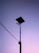 A silhouette of a tall street light with solar panels stands against a gradient sky transitioning from purple to pink. Electrical wires extend from the pole on the left side.