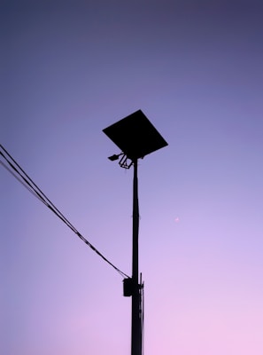 A silhouette of a tall street light with solar panels stands against a gradient sky transitioning from purple to pink. Electrical wires extend from the pole on the left side.