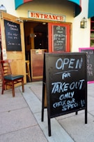 A restaurant entrance with open doors and chalkboard signs announcing margarita specials and takeout availability. The signs display operating hours, and a single chair is placed next to the open door. The building features a prominent 'Entrance' sign above the doorway.