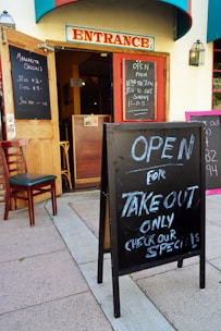 A restaurant entrance with open doors and chalkboard signs announcing margarita specials and takeout availability. The signs display operating hours, and a single chair is placed next to the open door. The building features a prominent 'Entrance' sign above the doorway.