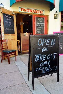 A restaurant entrance with open doors and chalkboard signs announcing margarita specials and takeout availability. The signs display operating hours, and a single chair is placed next to the open door. The building features a prominent 'Entrance' sign above the doorway.