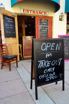 A restaurant entrance with open doors and chalkboard signs announcing margarita specials and takeout availability. The signs display operating hours, and a single chair is placed next to the open door. The building features a prominent 'Entrance' sign above the doorway.