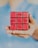 A hand holding a modified Rubik's Cube with red faces, each block displaying words. The words 'FIGURE IT OUT' are arranged in a sequence across the front of the cube. The background is softly blurred, highlighting the cube.