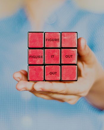 A hand holding a modified Rubik's Cube with red faces, each block displaying words. The words 'FIGURE IT OUT' are arranged in a sequence across the front of the cube. The background is softly blurred, highlighting the cube.