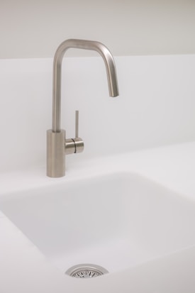 A modern, minimalist kitchen sink with a sleek, metallic faucet. The sink is white, and the faucet has a brushed metal finish, adding a contemporary touch. The kitchen setting appears clean and uncluttered, emphasizing simplicity and functionality.