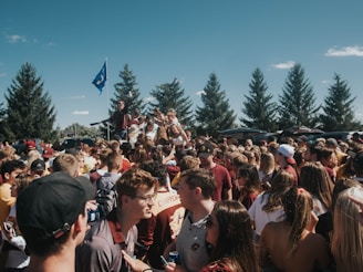 A large group of people gathered outdoors, likely at a party or event, with some individuals on a platform or vehicle holding a flag. The crowd is composed of mostly young adults, with many wearing casual clothing, and several individuals appear to be interacting and socializing. Tall trees are visible in the background along with a few cars, under a clear blue sky.