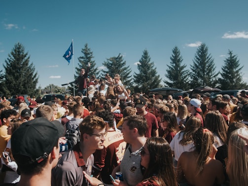 A large group of people gathered outdoors, likely at a party or event, with some individuals on a platform or vehicle holding a flag. The crowd is composed of mostly young adults, with many wearing casual clothing, and several individuals appear to be interacting and socializing. Tall trees are visible in the background along with a few cars, under a clear blue sky.