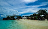 A tropical beach with clear turquoise water and soft white sand. Several lounge areas are set up under umbrellas and tents along the shoreline. Palm trees and lush greenery are visible in the background, with scattered clouds in a blue sky. People are seen enjoying the beach, swimming, and relaxing under the shaded areas.
