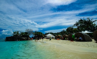 A tropical beach with clear turquoise water and soft white sand. Several lounge areas are set up under umbrellas and tents along the shoreline. Palm trees and lush greenery are visible in the background, with scattered clouds in a blue sky. People are seen enjoying the beach, swimming, and relaxing under the shaded areas.