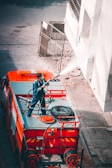 A person stands on top of a fire truck, directing a stream of water towards a building wall. The scene includes various equipment on the fire truck, such as hoses and a spare tire. The person is wearing a uniform, suggesting they are a firefighter. The image captures an urban environment with a partially visible gate and pavement.