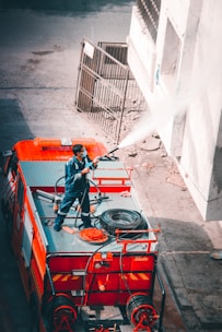 A person stands on top of a fire truck, directing a stream of water towards a building wall. The scene includes various equipment on the fire truck, such as hoses and a spare tire. The person is wearing a uniform, suggesting they are a firefighter. The image captures an urban environment with a partially visible gate and pavement.