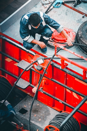 A person in a blue jumpsuit is engaged in work with various tools and equipment, crouching on a platform with industrial components. The platform and surrounding machinery are predominantly red with black hoses and pipes connected. The area appears to be an industrial or mechanical environment.