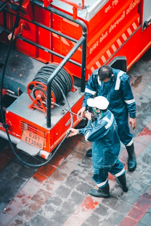 Two people dressed in blue protective gear are handling a hose connected to a red vehicle, possibly a fire truck. The ground is paved with gray and red tiles, and the vehicle has writing in a language other than English, possibly indicating it