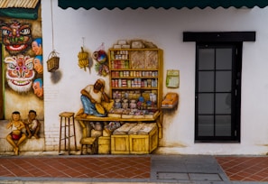 A vibrant mural depicting a small, colorful street scene. On the left, a wall features traditional, folkloric masks and three playful children sitting against it. There is a shopkeeper resting on a wooden counter surrounded by various goods like snacks, household items, and newspapers displayed neatly. The background includes a black-framed glass door and an awning overhead.