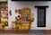A vibrant mural depicting a small, colorful street scene. On the left, a wall features traditional, folkloric masks and three playful children sitting against it. There is a shopkeeper resting on a wooden counter surrounded by various goods like snacks, household items, and newspapers displayed neatly. The background includes a black-framed glass door and an awning overhead.