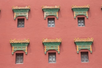 A series of windows with ornate, decorative architectural elements line a red wall. The windows have intricate lattice designs, and above them are detailed cornices painted in green and orange, accented with gold.
