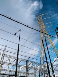 High voltage electrical transmission towers and power lines dominate the scene against a partly cloudy blue sky. Various metal structures and a streetlight are also visible, forming part of an electrical substation.