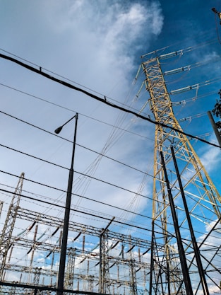 High voltage electrical transmission towers and power lines dominate the scene against a partly cloudy blue sky. Various metal structures and a streetlight are also visible, forming part of an electrical substation.