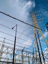 High voltage electrical transmission towers and power lines dominate the scene against a partly cloudy blue sky. Various metal structures and a streetlight are also visible, forming part of an electrical substation.