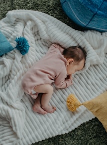 A baby is sleeping peacefully on a soft, white, ribbed blanket with a cozy atmosphere. The baby is wearing a light pink ribbed onesie. Around the baby, there are cushions in shades of blue and a small yellow pom-pom for decoration on the blanket. The background includes a textured green carpet.