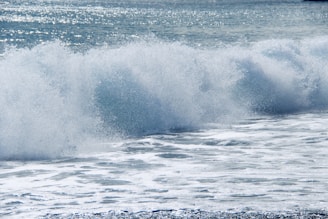 A large ocean wave crashing with force, creating white foam and spray. The water is a mix of blue and white, glistening under sunlight. The sea extends into the distance with a sparkling surface.