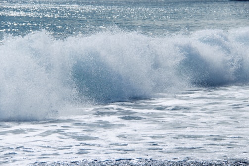 A large ocean wave crashing with force, creating white foam and spray. The water is a mix of blue and white, glistening under sunlight. The sea extends into the distance with a sparkling surface.