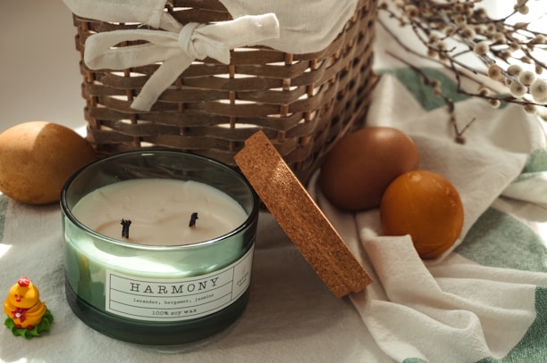 A wicker basket with a fabric cover is situated among various decorative items. A glass jar candle labeled 'Harmony' with notes of lavender, bergamot, and jasmine is in the foreground, its lid resting beside it. Two rustic brown eggs and a small ceramic figurine of a yellow bird wearing a floral wreath are included in the scene. The background features cotton branches resting on a soft, striped cloth.