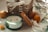 A wicker basket with a fabric cover is situated among various decorative items. A glass jar candle labeled 'Harmony' with notes of lavender, bergamot, and jasmine is in the foreground, its lid resting beside it. Two rustic brown eggs and a small ceramic figurine of a yellow bird wearing a floral wreath are included in the scene. The background features cotton branches resting on a soft, striped cloth.