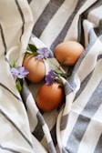 Three brown eggs nestled on a striped fabric with delicate purple flowers and green leaves intertwined around them.