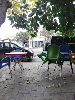 A street scene with plastic chairs stacked on a tiled pavement, colorful in green, blue, and yellow. A table with a red top is also visible. Leafy branches hang overhead, while a black car is parked nearby. The background features a sidewalk, palm trees, and a building with glass windows.